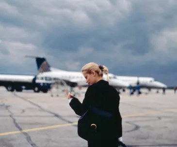 Person with a backpack walking on an airport tarmac near a small airplane under cloudy skies.