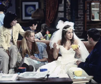 Group of friends on a coffeehouse couch; woman in wedding dress holding a yellow mug, discussing intently.