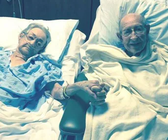 Elderly couple in hospital beds holding hands, wrapped in blankets, looking towards the camera.
