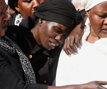 A woman in a black headscarf leans on another for support, both wearing solemn expressions.
