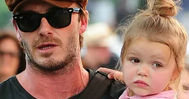 A man wearing sunglasses and a cap holds a young child with a pacifier in her mouth.