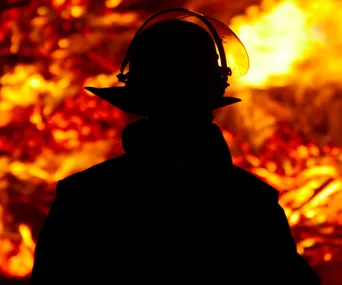 A firefighter silhouetted against large flames, wearing a helmet and protective gear.