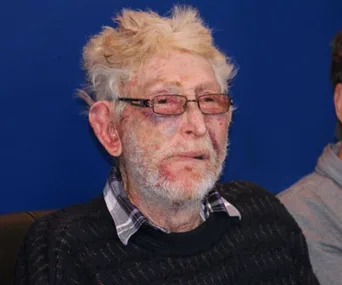 Elderly man with glasses, visible bruises and injuries on his face, wearing a sweater and checkered shirt, against a blue background.