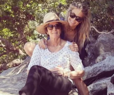 Two women outdoors on a rocky terrain; one sitting with a sun hat and sunglasses, the other standing, smiling, in sunglasses.