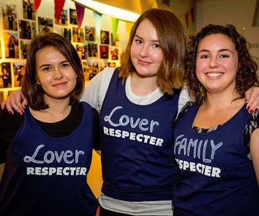 Three smiling women wearing shirts labeled "Lover Respected" and "Family Respected" pose together indoors.