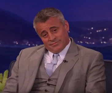Matt LeBlanc in a suit, sitting and smirking during an interview, with a blurred night cityscape background.