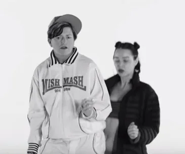 Two individuals dancing; the front person wears a cap and jacket with "Mish Mash" text. Black and white image.