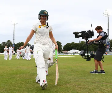 Australian women's cricket player leaving the field, followed by teammates and a cameraman.
