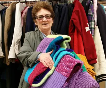 Woman holding colorful knitted jumpers stands in front of a rack of coats.