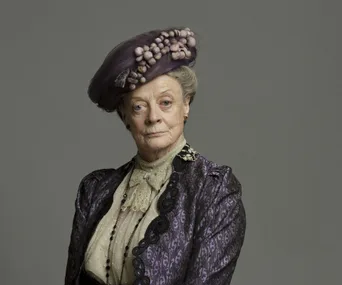 Dame Maggie Smith as Violet Crawley in "Downton Abbey," wearing a vintage purple outfit with an elaborate hat.