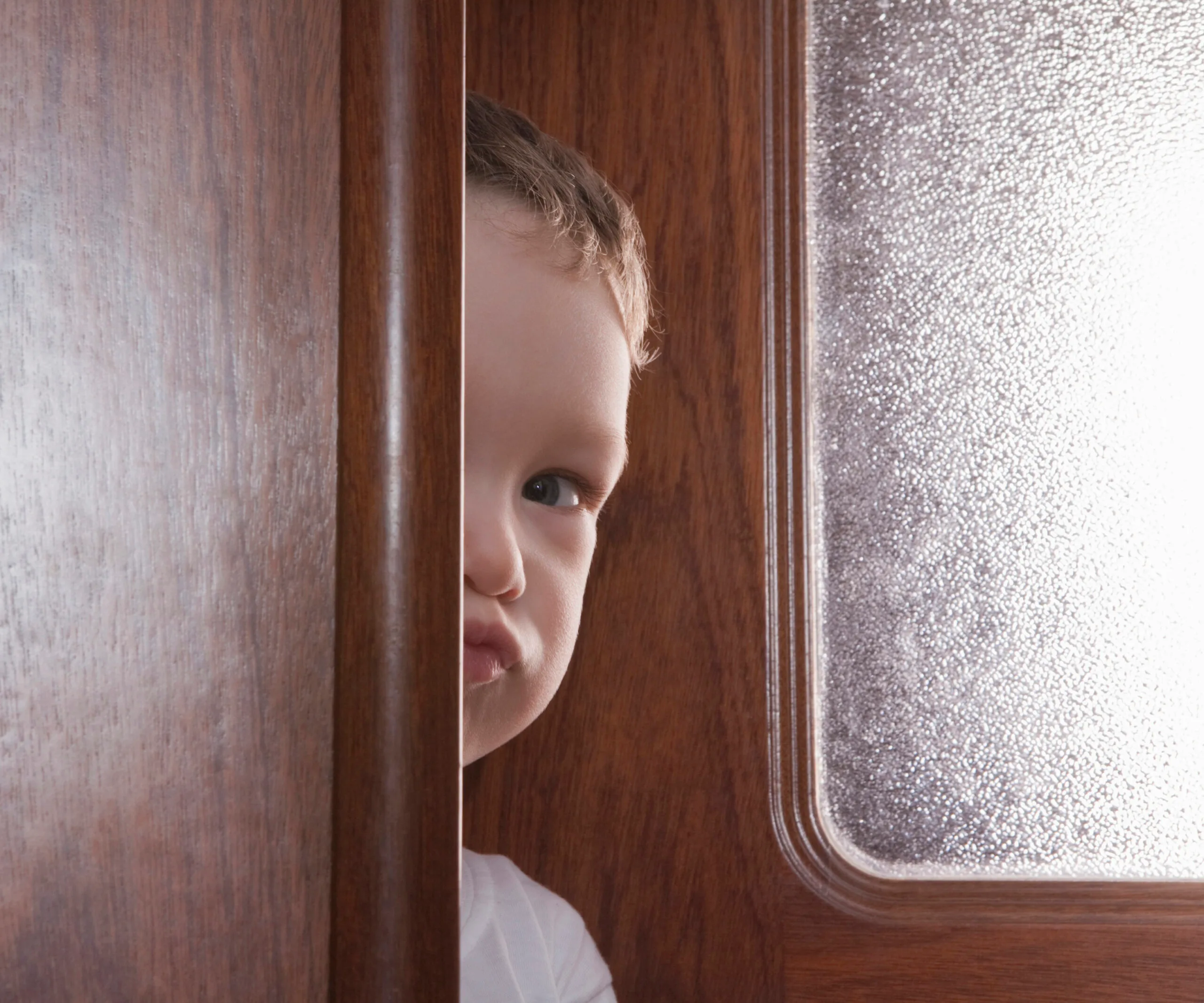 A young boy peeks through a partially open wooden door with frosted glass, only half his face visible.