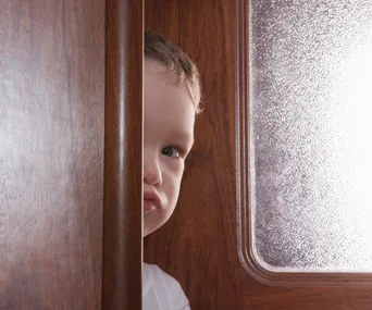 A young boy peeks through a partially open wooden door with frosted glass, only half his face visible.