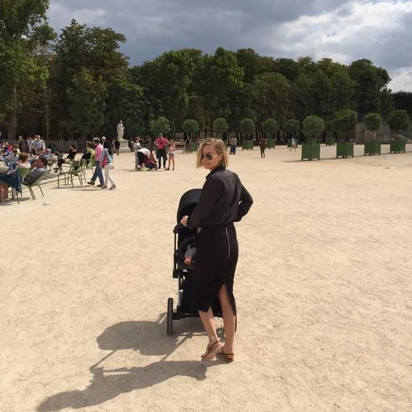 A woman in sunglasses pushes a stroller in a sunny Paris park, surrounded by trees and people.