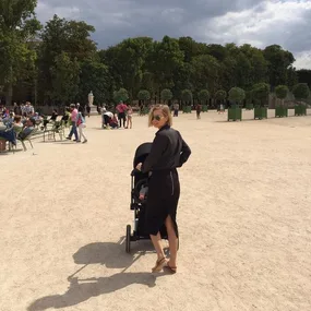 A woman in sunglasses pushes a stroller in a sunny Paris park, surrounded by trees and people.