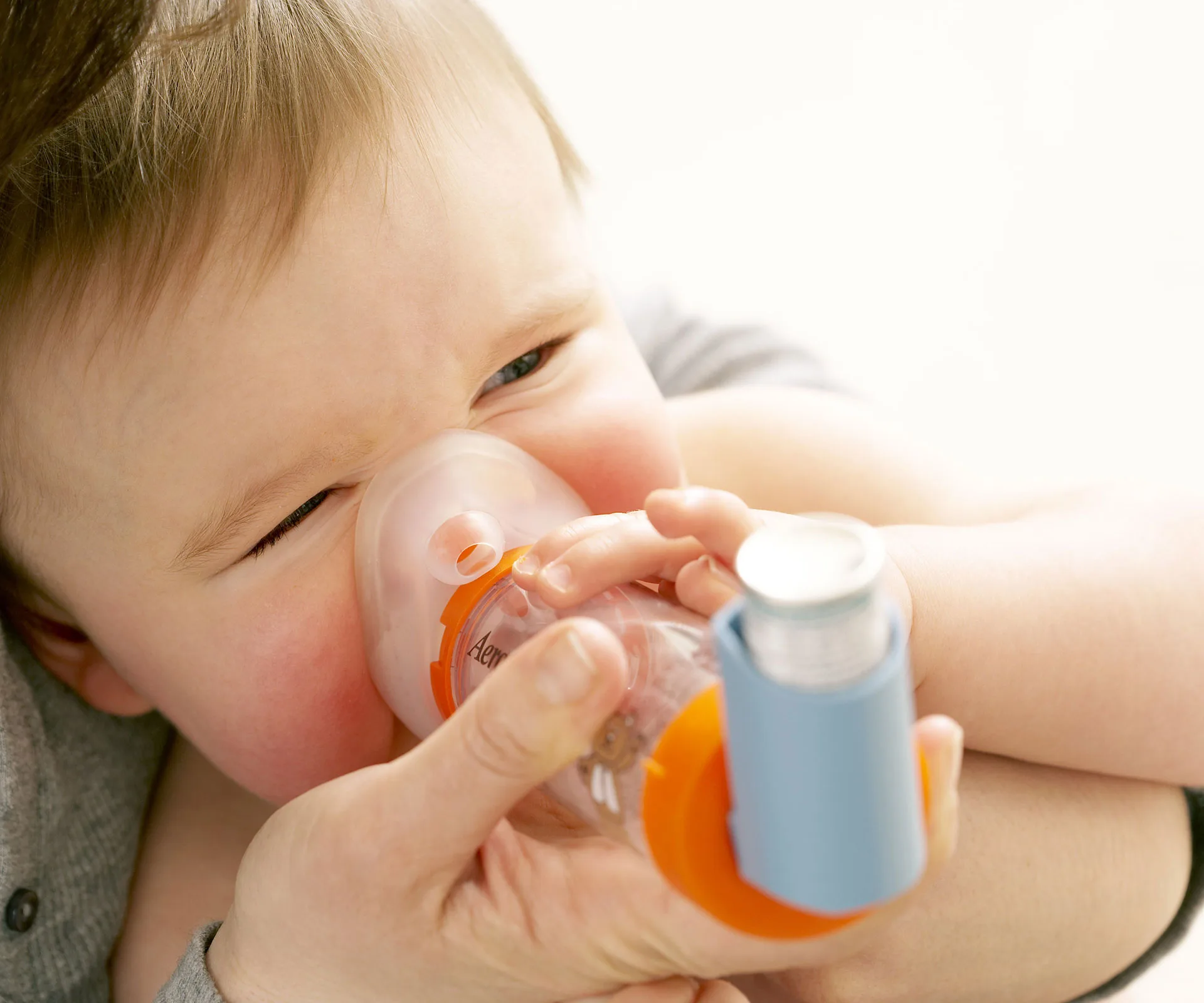 Baby using a nebulizer mask, assisted by an adult, for asthma treatment.