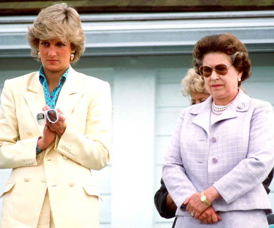 Two women stand side by side; one in a yellow suit, holding sunglasses, the other in a checked lavender suit, both outdoors.