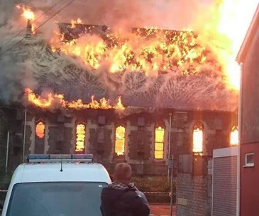Church engulfed in flames, person in foreground taking photo, emergency vehicle present.