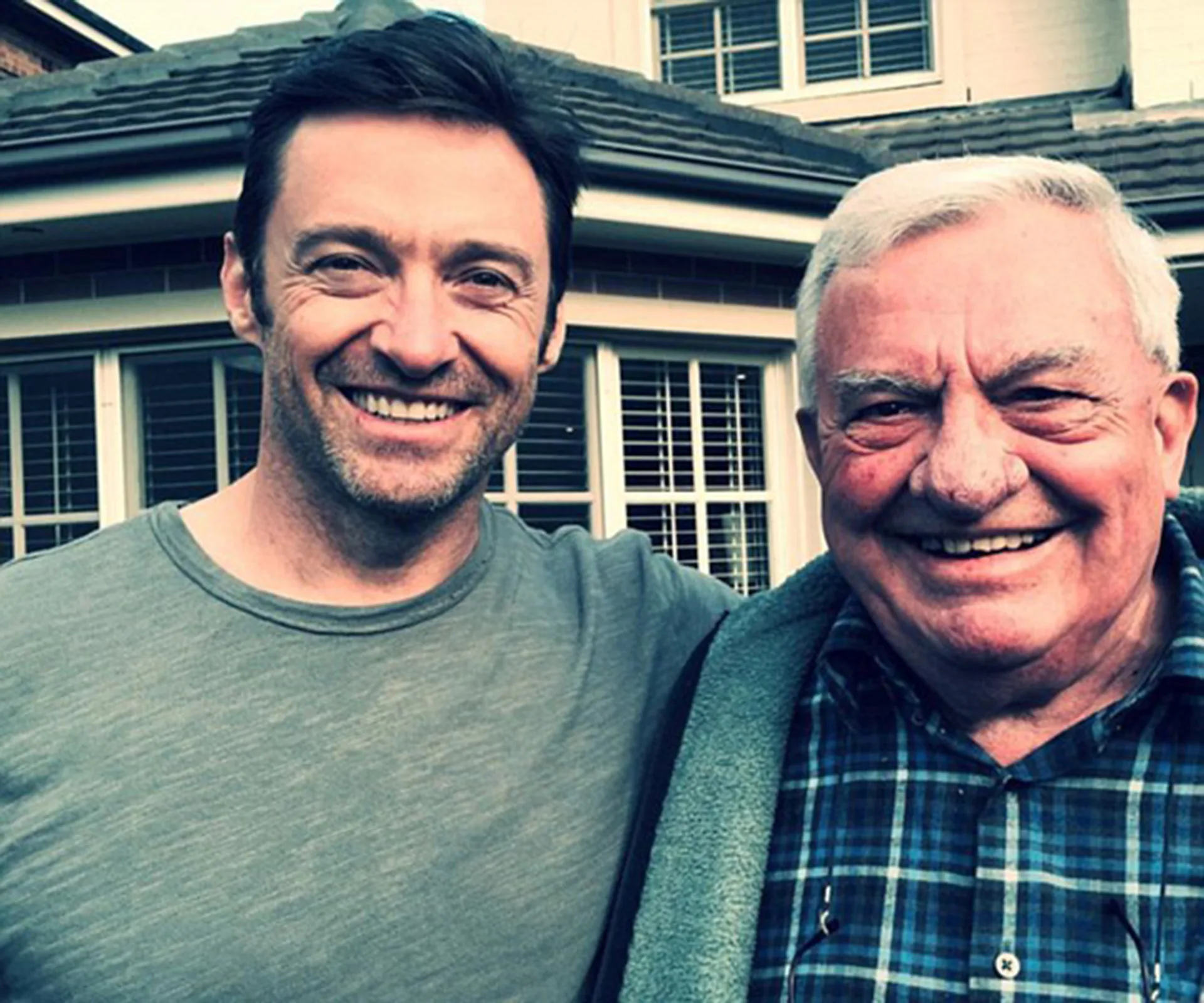 Smiling man with short dark hair beside an older man with white hair, both outdoors in front of a house.