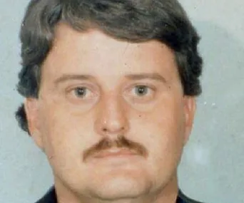 A close-up of a man with brown hair, mustache, wearing a dark top, neutral expression, against a plain background.