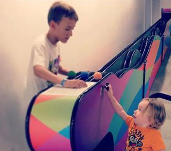Two young children play at a colorful, interactive wall with balls, one standing and one reaching up.