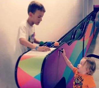 Two young children play at a colorful, interactive wall with balls, one standing and one reaching up.