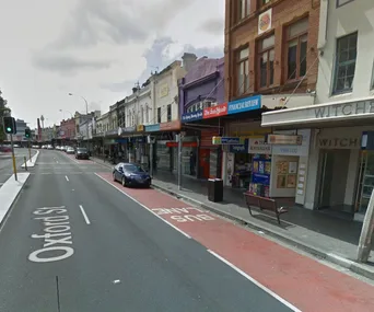 Street view of Oxford Street in Sydney, showcasing shops with signs and a bus lane beside a main road.