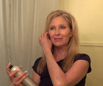 A woman in a black shirt styles her long blonde hair with hairspray in front of a yellow bathroom wall.