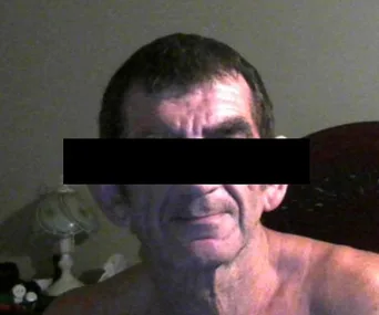 A man with his eyes obscured by a black bar, sitting shirtless in a dim room with a lamp in the background.