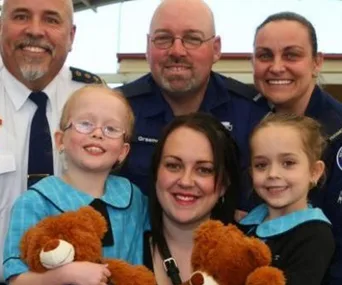 Group of people smiling, including children in blue uniforms holding teddy bears, and adults in uniforms in the background.