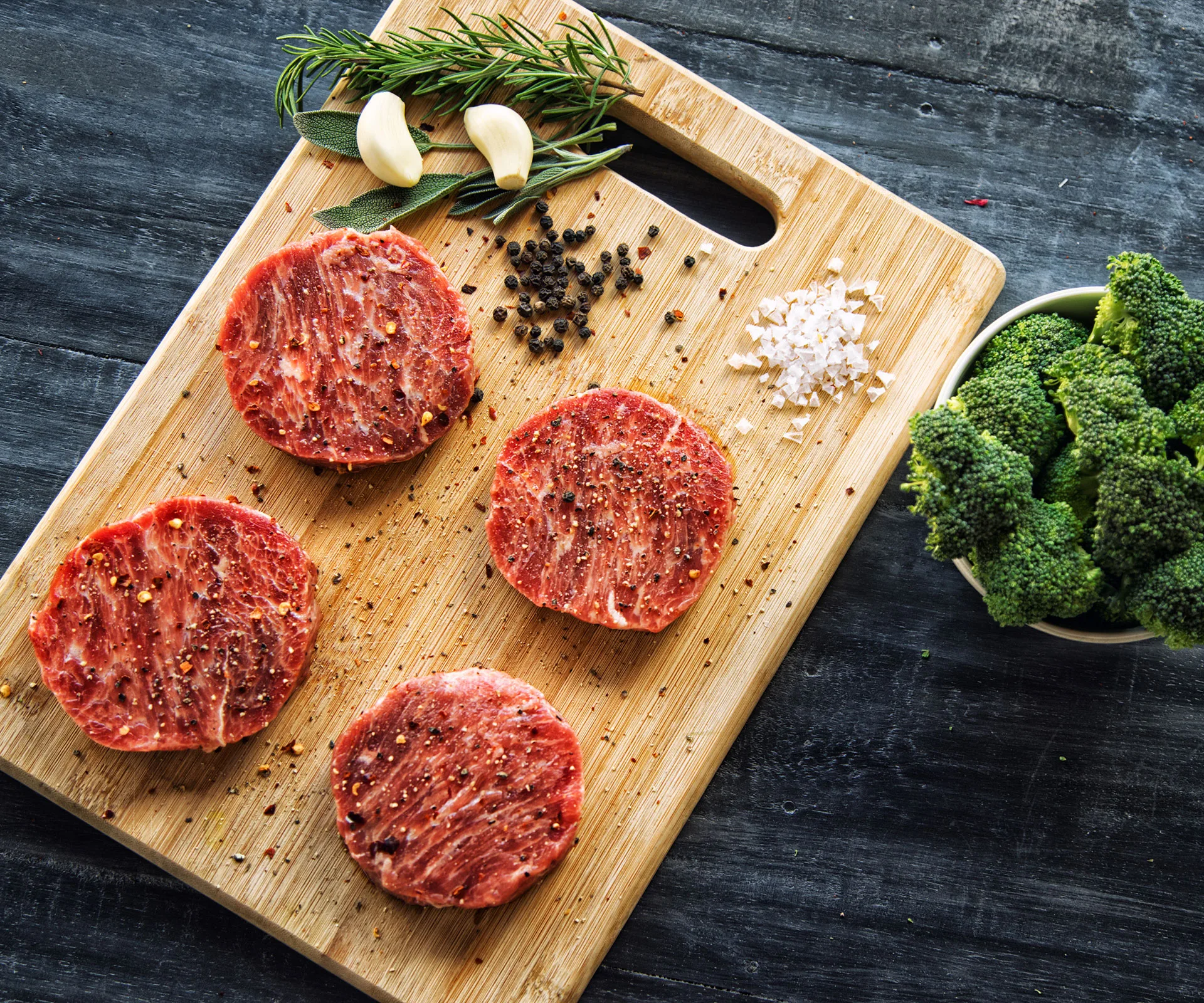 Raw beef rounds seasoned with spices on a wooden board, with garlic, herbs, and broccoli nearby.