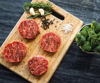 Raw beef rounds seasoned with spices on a wooden board, with garlic, herbs, and broccoli nearby.