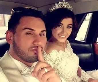 Man in a tuxedo and woman in a wedding dress with a tiara, smiling in a car. Man gestures for silence with his finger.