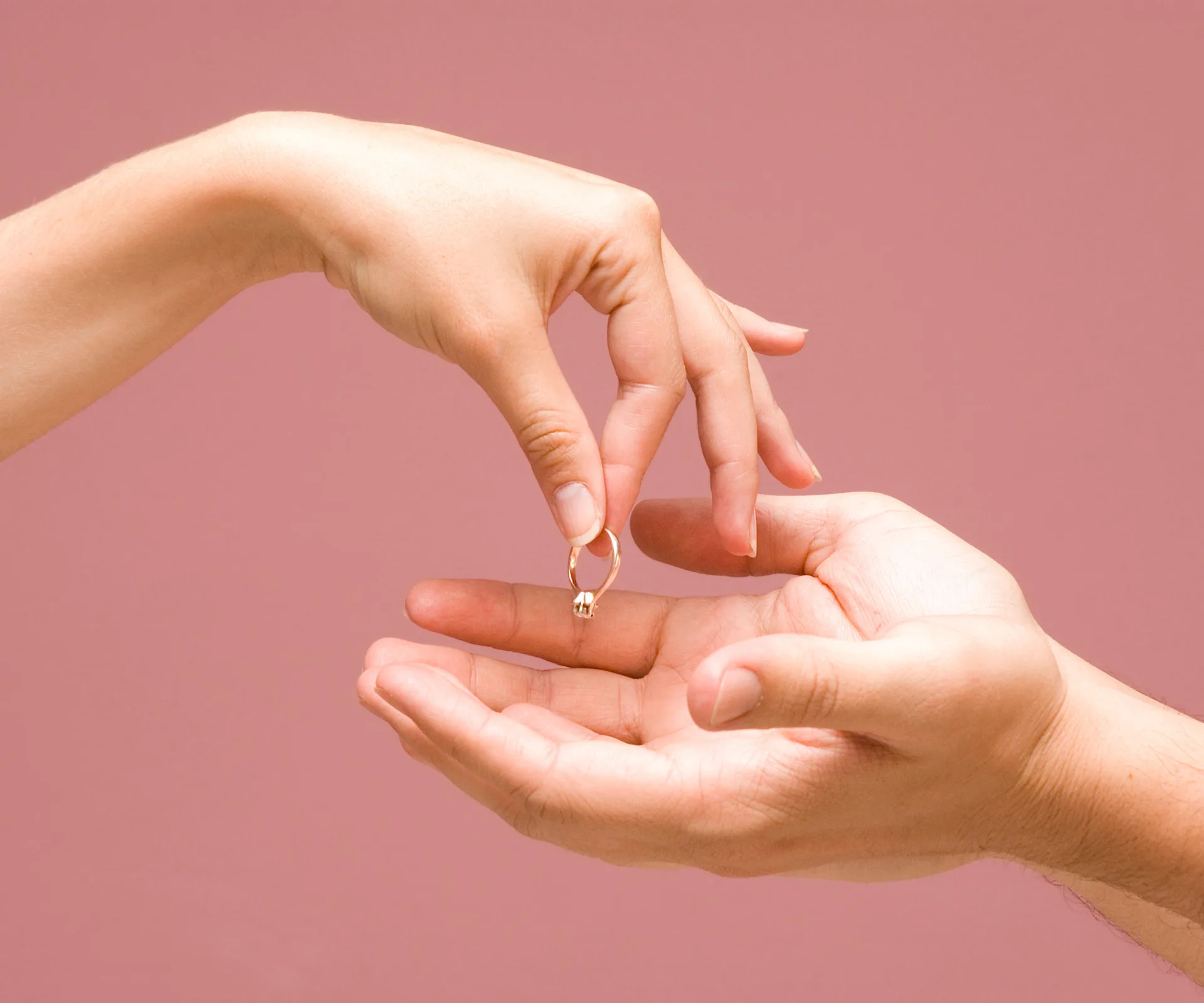 Hands exchanging a ring, set against a pink background, symbolizing a breakup or returned engagement.