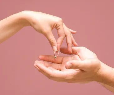 Hands exchanging a ring, set against a pink background, symbolizing a breakup or returned engagement.