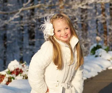 Girl in white coat and flower headband smiling outdoors in snowy scenery.