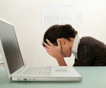 Woman sitting at a desk with head in hands, facing a laptop, appearing stressed or frustrated.