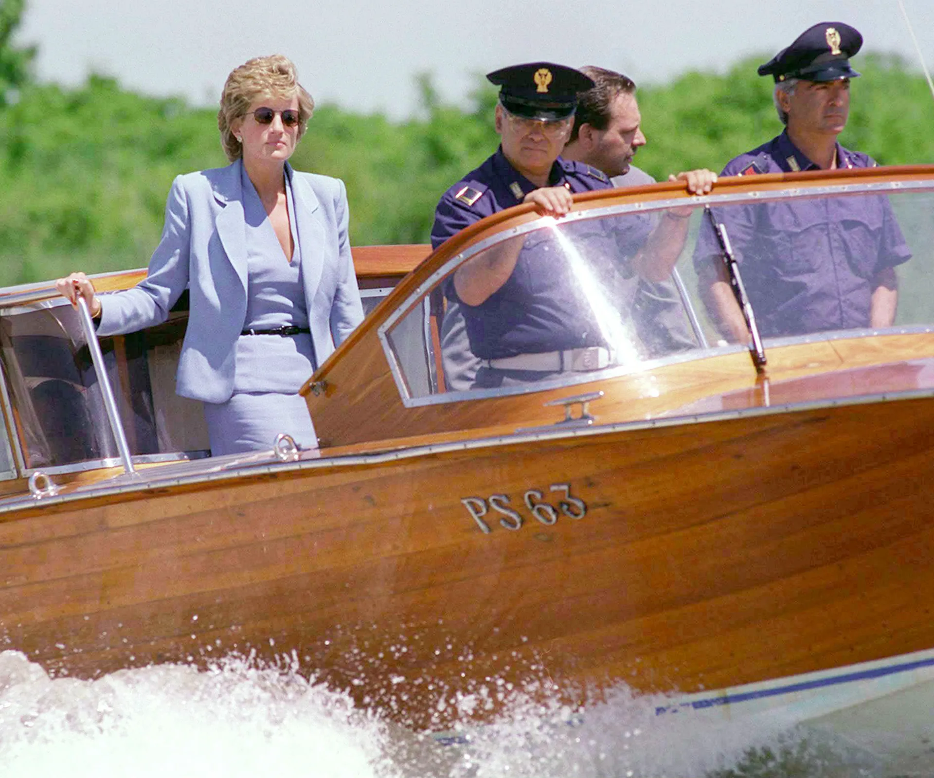 Woman in a blue suit on a wooden boat with uniformed men, navigating through water.