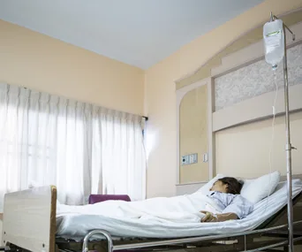A person lying in a hospital bed with an IV, in a softly lit room with light-colored curtains.