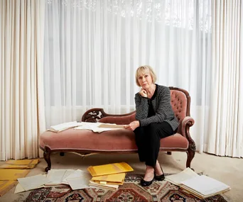 Elderly woman seated on vintage sofa, surrounded by scattered papers and folders, looks contemplative near sheer curtains.
