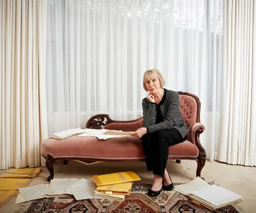 Elderly woman seated on vintage sofa, surrounded by scattered papers and folders, looks contemplative near sheer curtains.