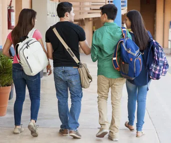 Four students with backpacks walk together down a school hallway, discussing.