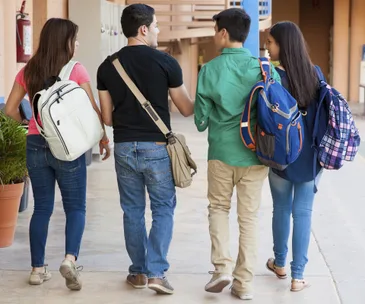 Four students with backpacks walk together down a school hallway, discussing.