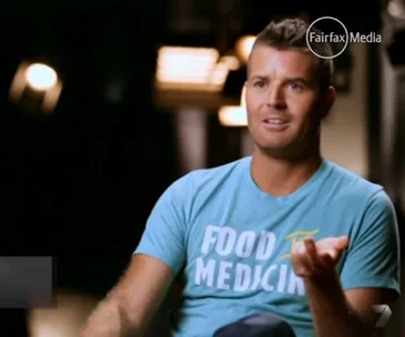 Man in blue "Food is Medicine" shirt, gesturing, sitting indoors.