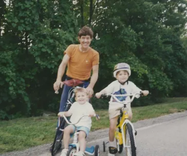 A smiling man with two young boys on bicycles, one in a helmet, on a tree-lined road.
