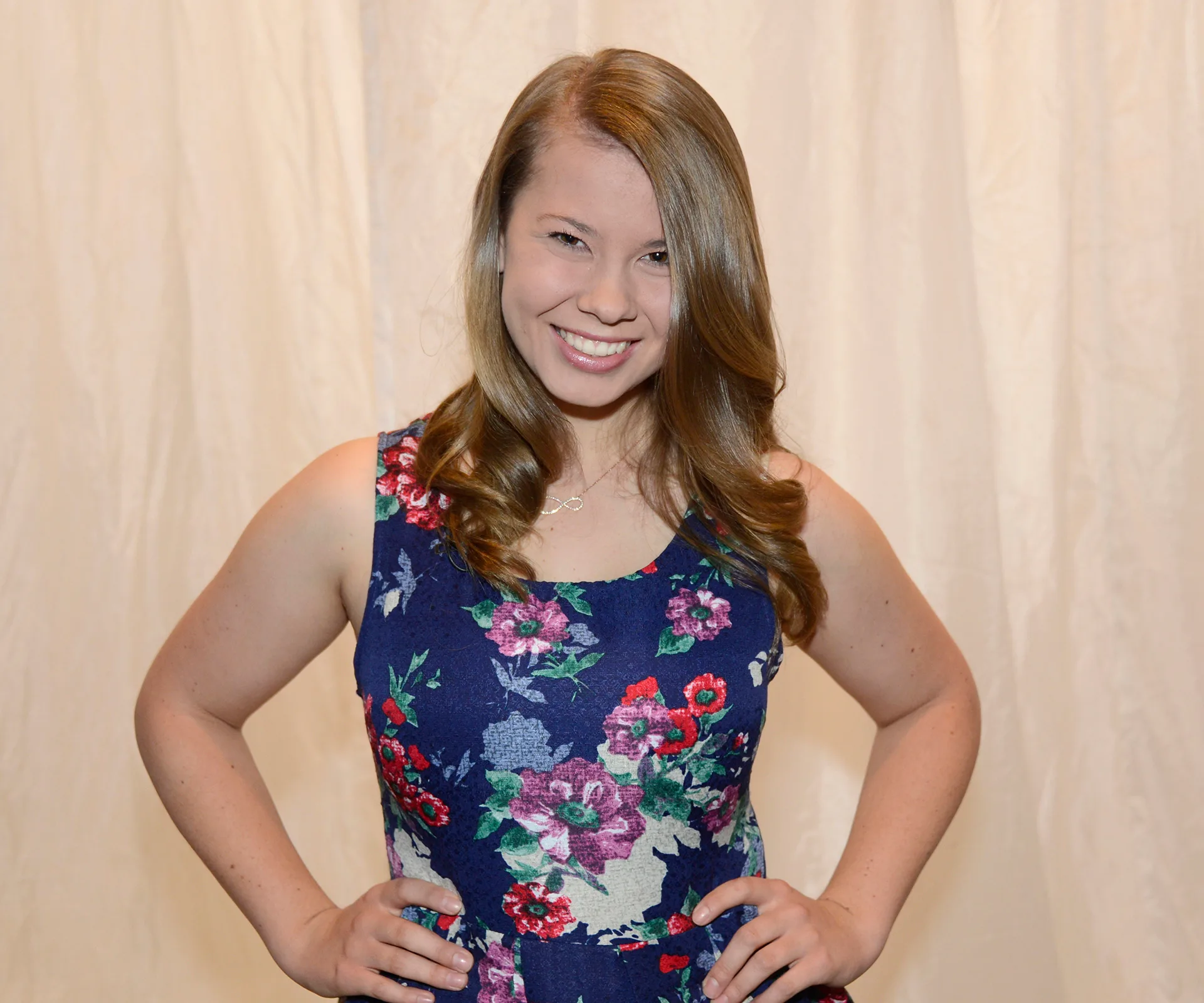 Young woman smiling in a floral dress, hands on hips, against a light curtain background.