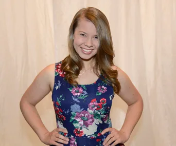 Young woman smiling in a floral dress, hands on hips, against a light curtain background.