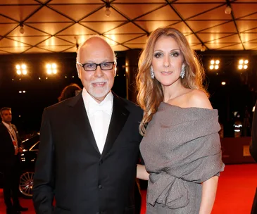 A woman in an elegant dress stands beside a man in a tuxedo on a red carpet with lights in the background.