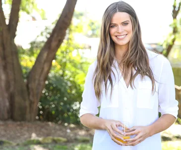 A person in a white shirt smiles holding a glass of juice in a sunny outdoor setting.