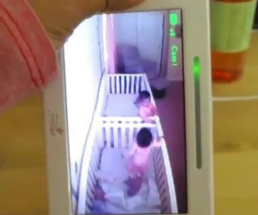 Two babies seen on a monitor standing and moving in their cribs, appearing to be alert.