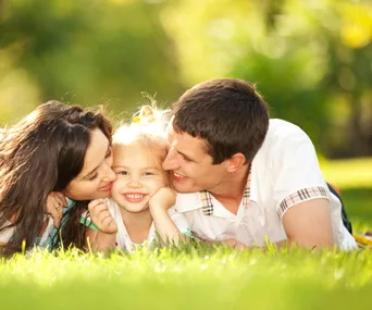 A family of three, a mother, father, and child, smiling and lying on the grass in a sunny, green park.